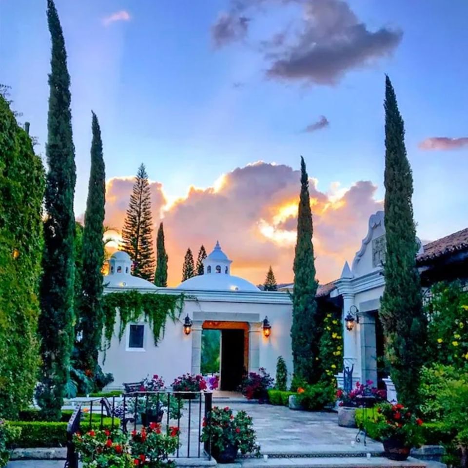 Patio de piedra con árboles altos rodeando cúpulas blancas bajo un cielo de atardecer colorido en Pensativo House Hotel