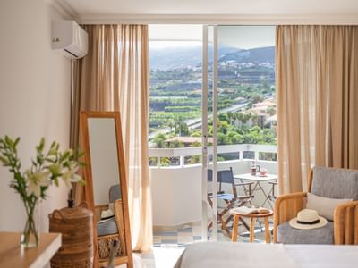 A hotel room with balcony and scenic view of a valley and buildings, featuring a bed, chairs, and table.