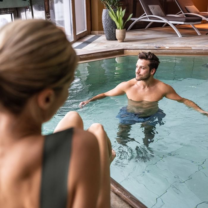 Uomo in piscina con donna seduta a bordo, in una spa con piante e lettini.