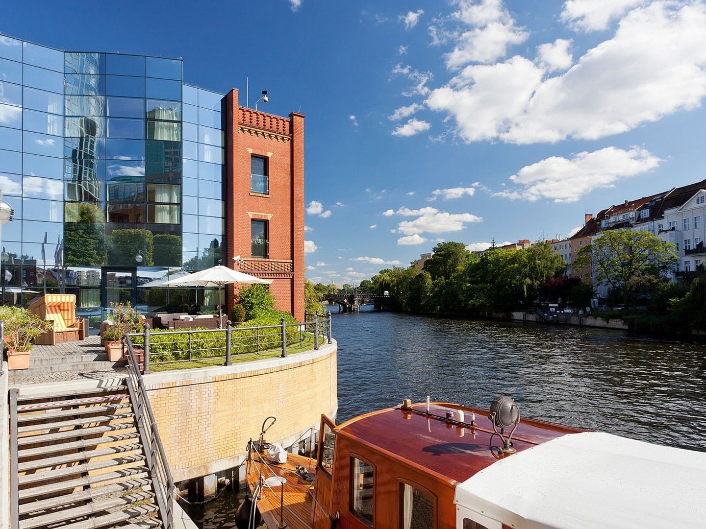 Modernes Gebäude mit Glasfassaden, Booten und Treppen an einem Fluss mit Bäumen und blauem Himmel.
