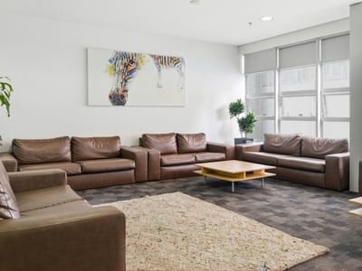 A cozy lounge area with brown couches, wooden tables, and a large painting at Student Living Auckland – Anzac.