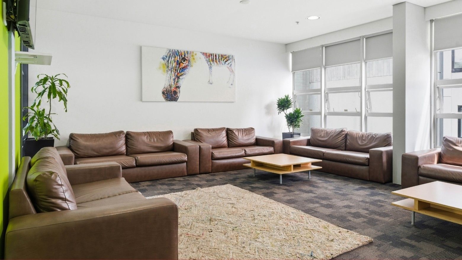 A cozy lounge area with brown couches, wooden tables, and a large painting at Student Living Auckland – Anzac.