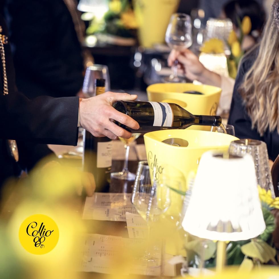Hand pouring Collio wine into glass with yellow buckets and flowers at an event.