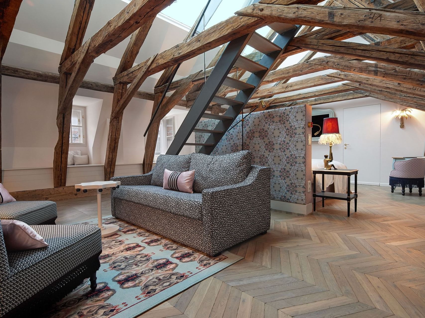 A spacious hotel suite with exposed wooden beams, patterned furniture, a staircase, and a skylight.