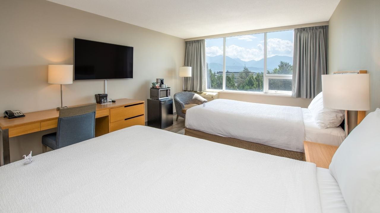 Hotel room with two beds, desk, chair, TV, lamp, and large window with mountain view.
