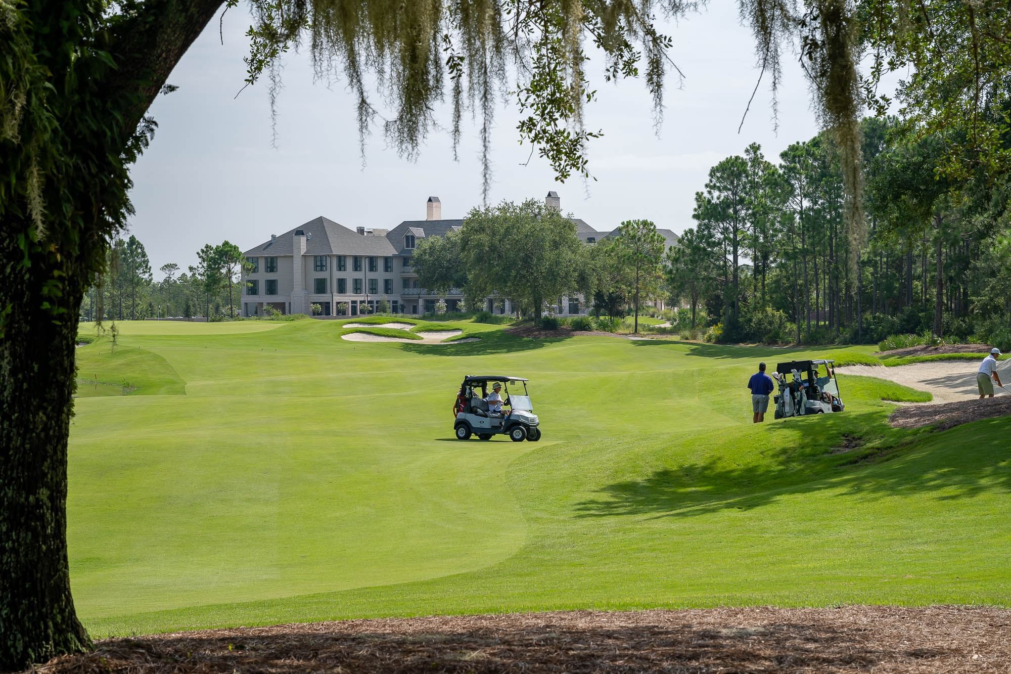 Golfers in carts on a green golf course with a large building in the background and trees around.