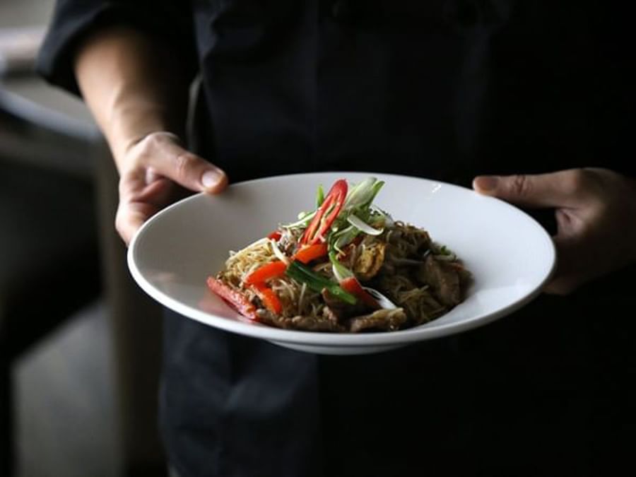 Stir-fried noodles with meat and vegetables held in a white bowl by a chef at Warwick Le Crystal Montreal