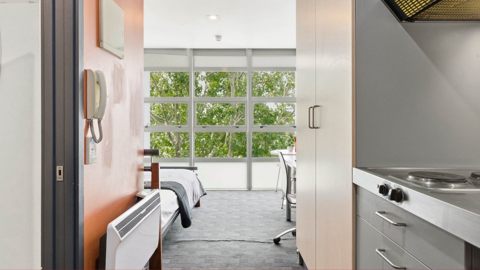 Compact bedroom with bed, desk, and kitchenette in a student living space.