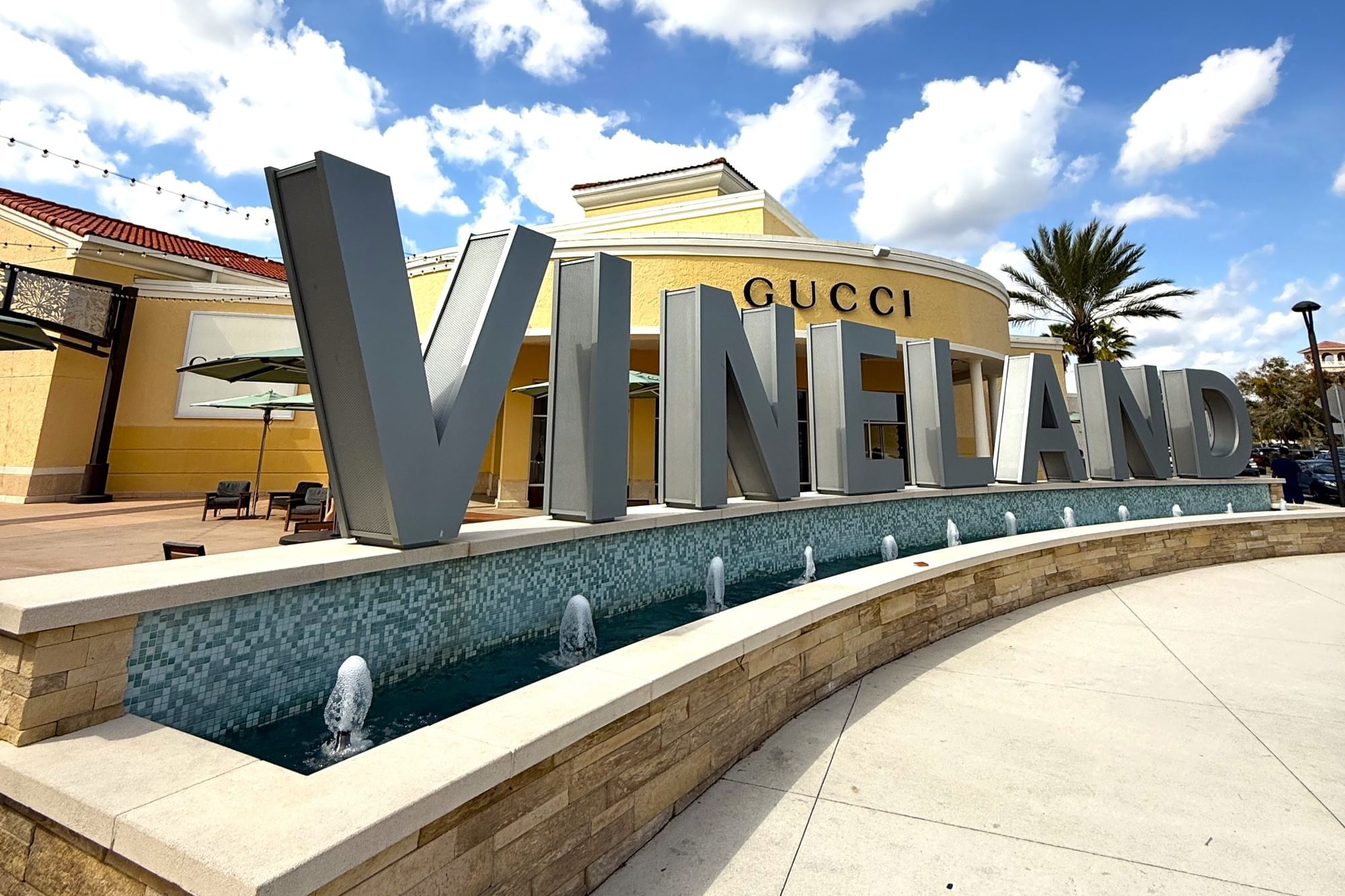 A large sign reading Vineland with a decorative fountain curving in front of a tan stucco building with a sign that reads Gucci.