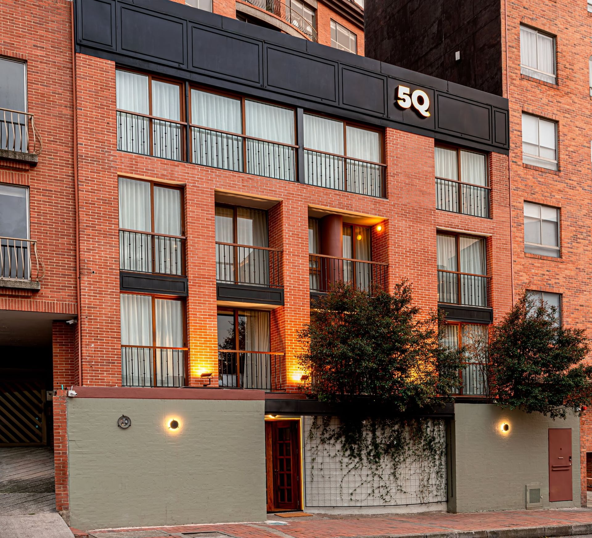 Red brick exterior of 5Q House Chapinero with black window frames by green trees under a glowing hotel name sign