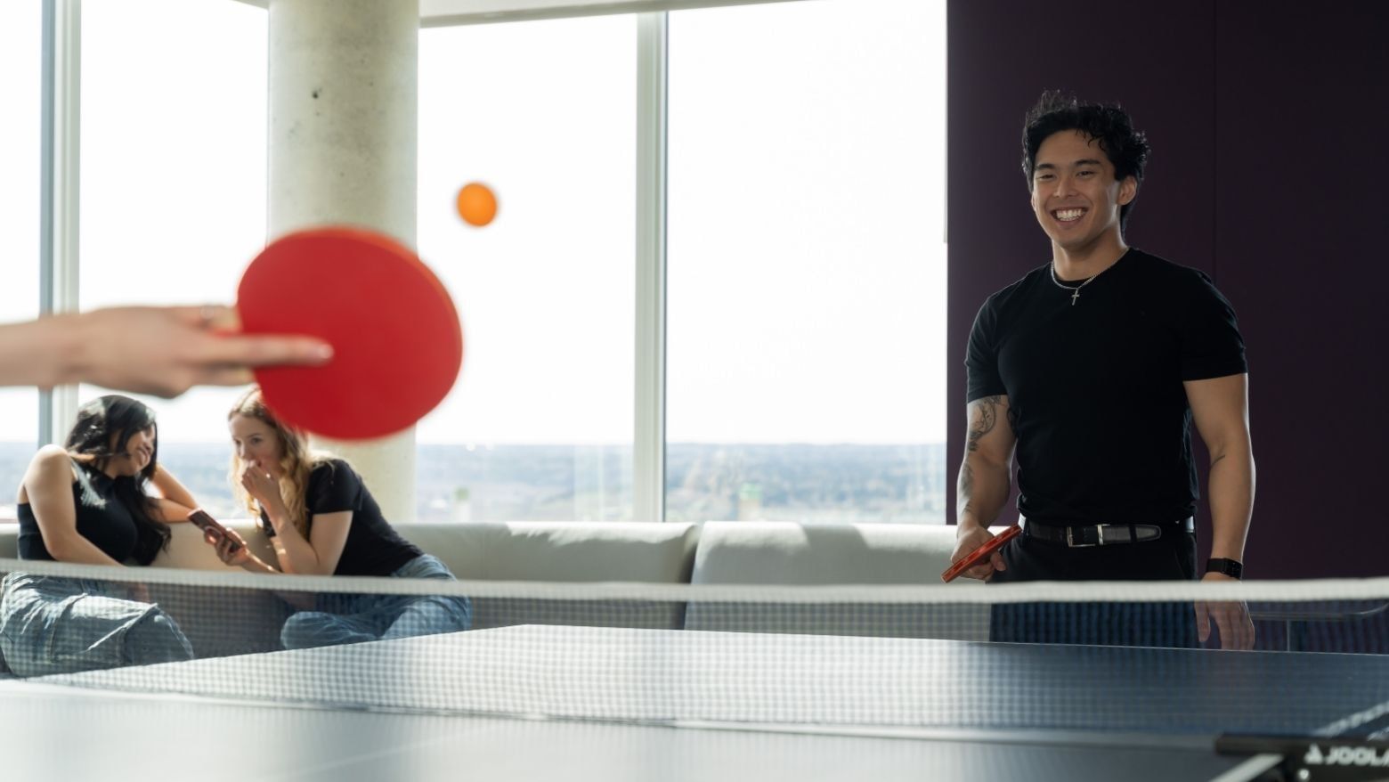Students playing ping pong and chatting on the lounge chairs, with a view of the sky from floor to ceiling windows.