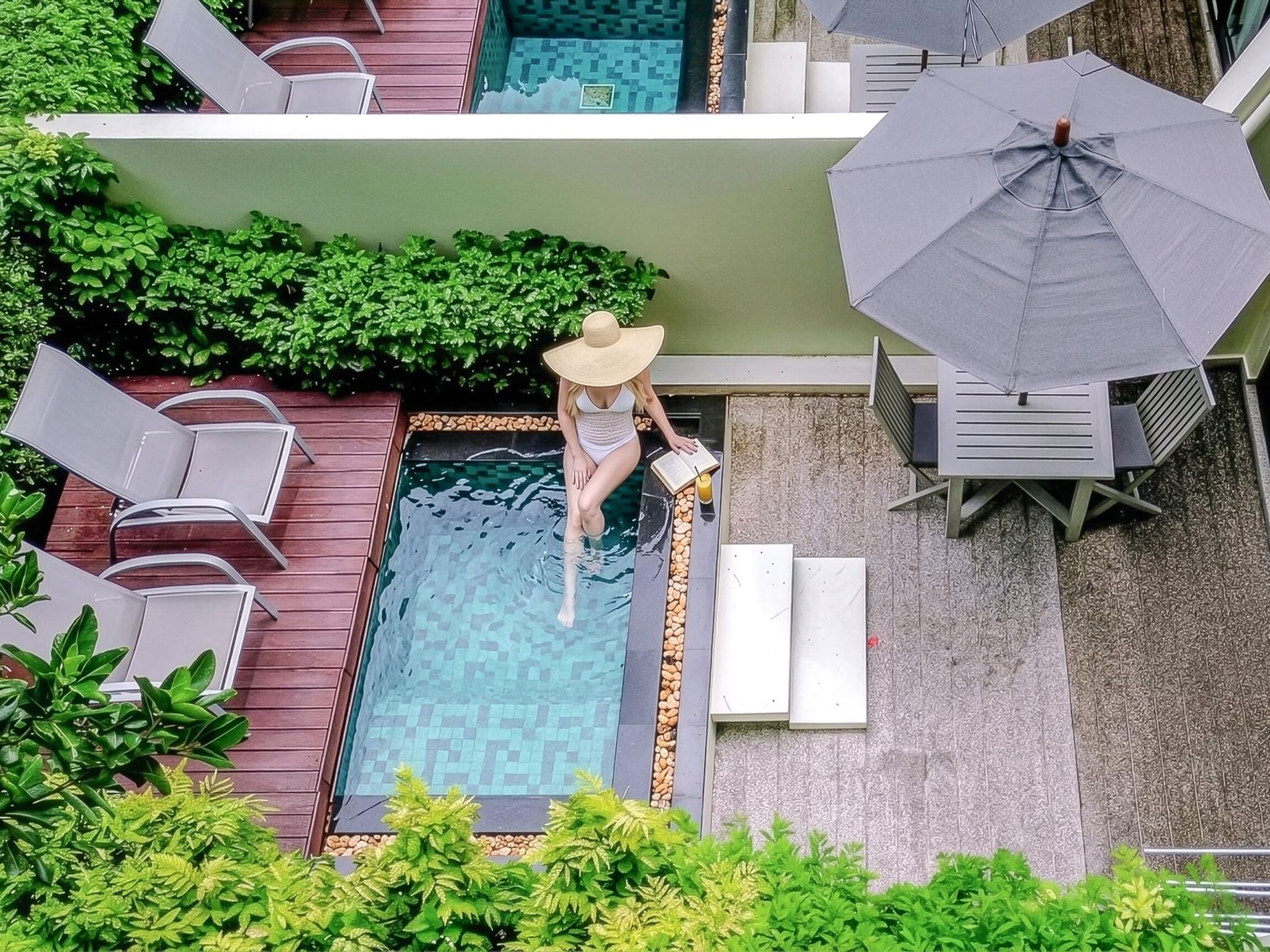 Aerial view of woman in sun hat relaxing in private plunge pool surrounded by tropical greenery and modern patio furniture.