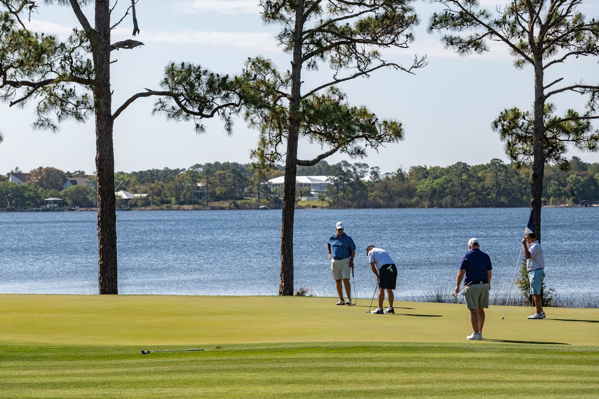 Four golfers with clubs play by a lake with trees and distant houses.