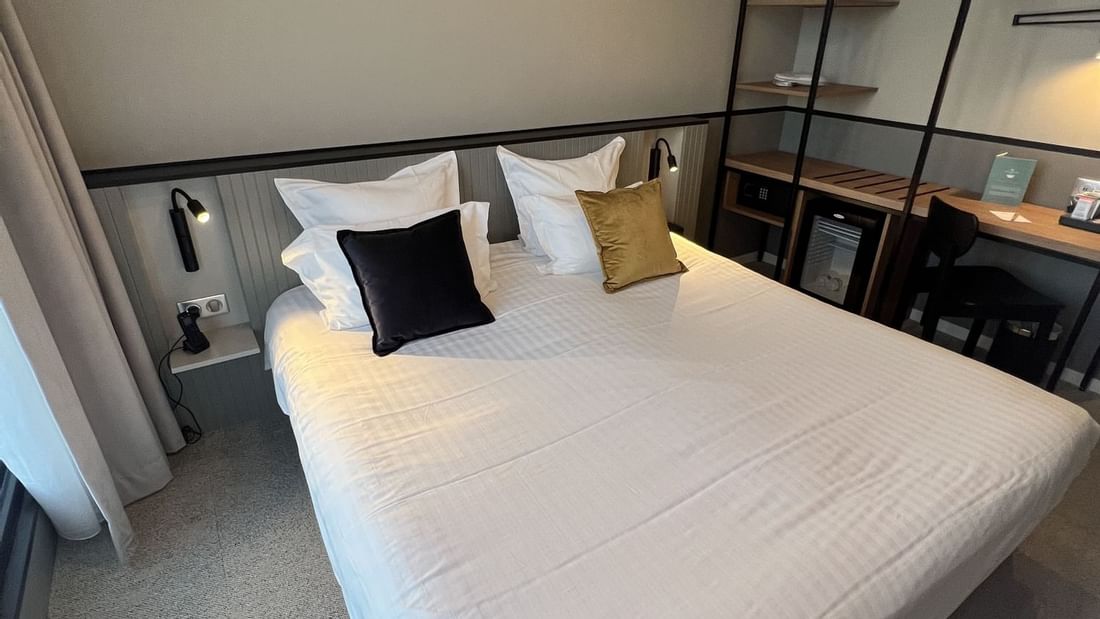 A clean hotel room with a large bed, nightstand, shelf, desk, and hanging light fixture.