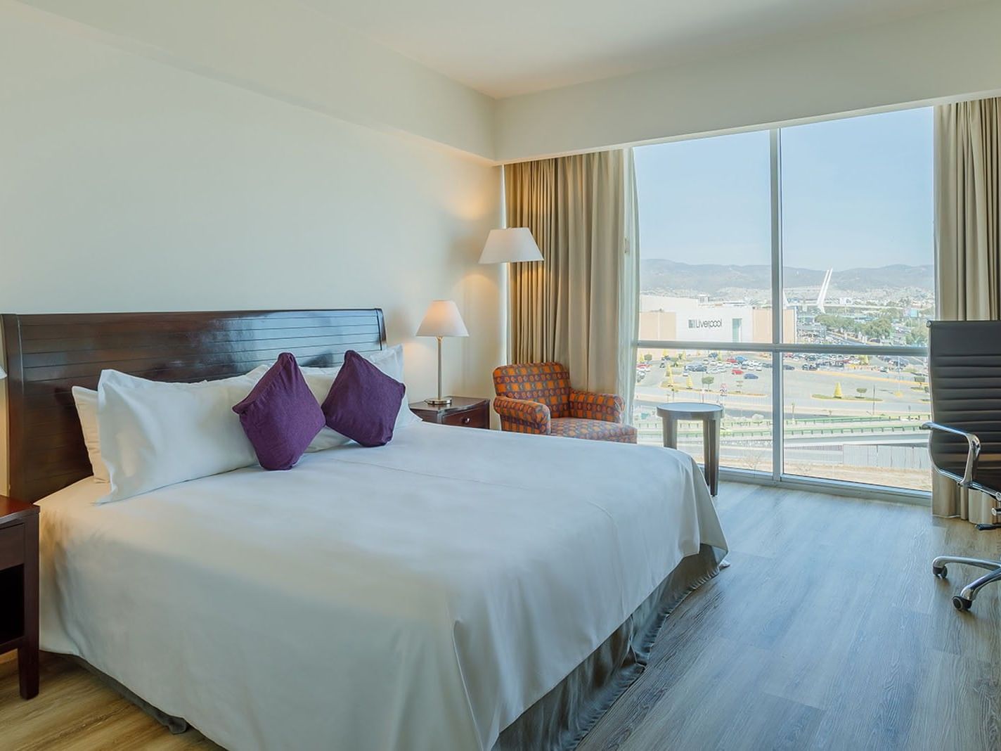 King Deluxe room featuring purple pillows, a patterned armchair, and a wide city window view at Camino Real Pachuca