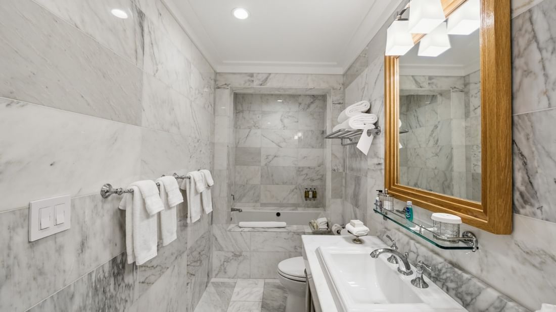 Marble bathroom with tub, white towels, wood-framed mirror, and glass shelf in Deluxe King Suite at el PRADO Hotel.