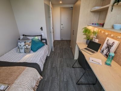 Modern twin bedroom with light wood desk, laptop, cork board, and teal accent pillows in Tupuārangi.
