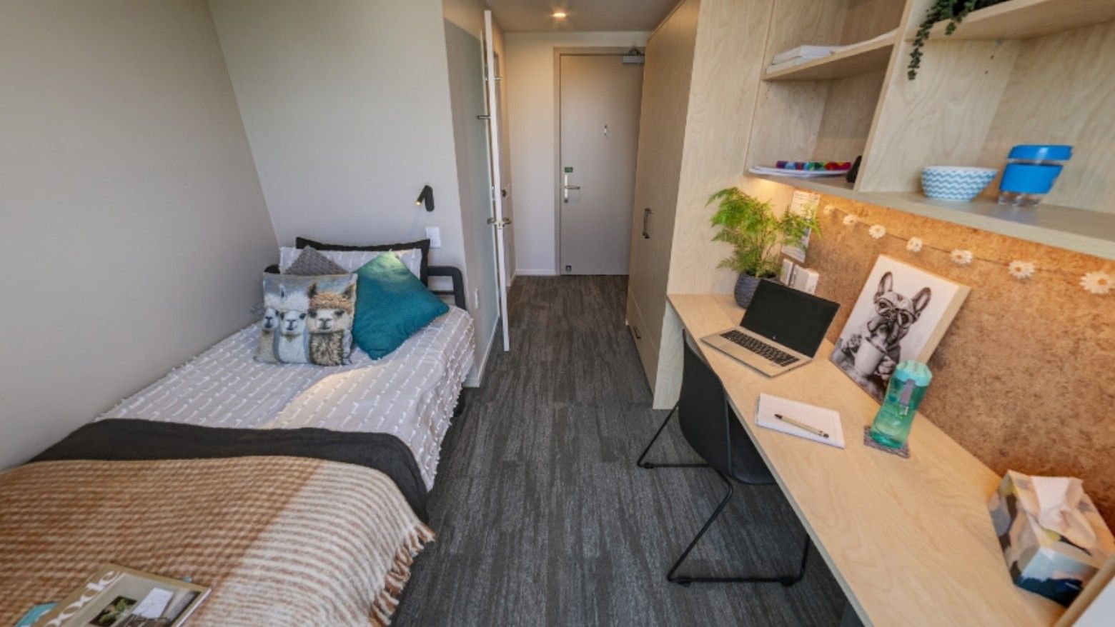 Modern twin bedroom with light wood desk, laptop, cork board, and teal accent pillows in Tupuārangi.