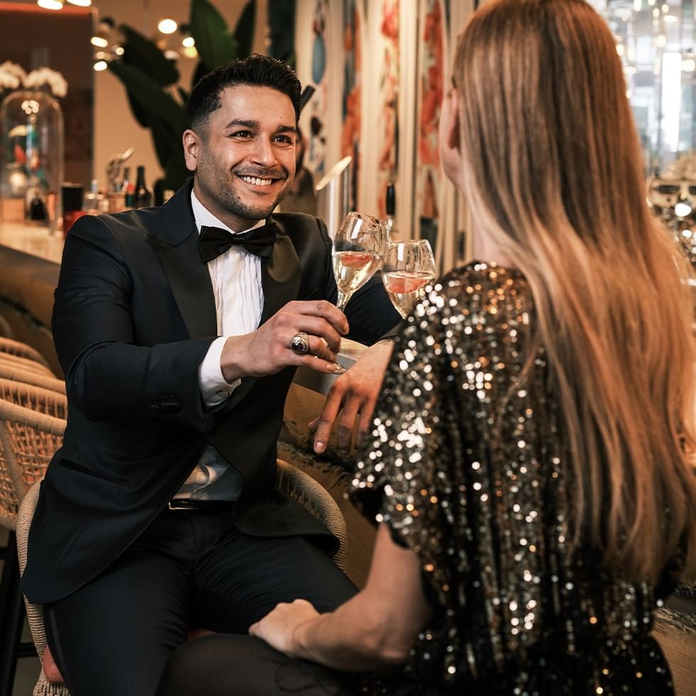 Uomo in smoking e donna in abito scintillante brindano con champagne in un elegante bar.