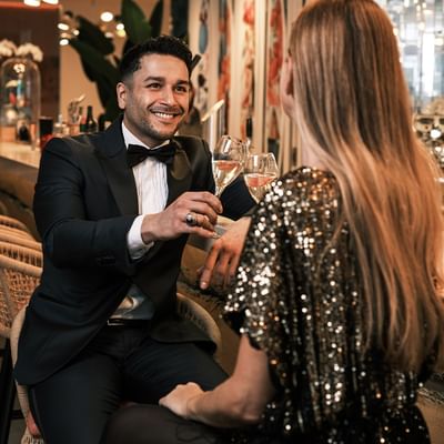 Uomo in smoking e donna in abito scintillante brindano con champagne in un elegante bar.