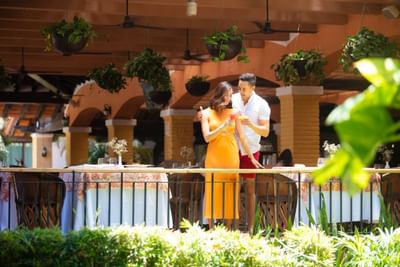Couple standing together on a balcony, holding drinks, with tables and hanging plants in the background.