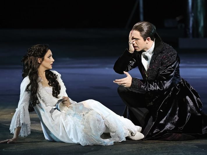 A man and woman in period costumes sit on a stage, the man kneeling with his hand on his face.