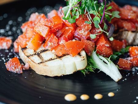 A black plate with a dish containing a slice of bread topped with diced tomatoes and greens.
