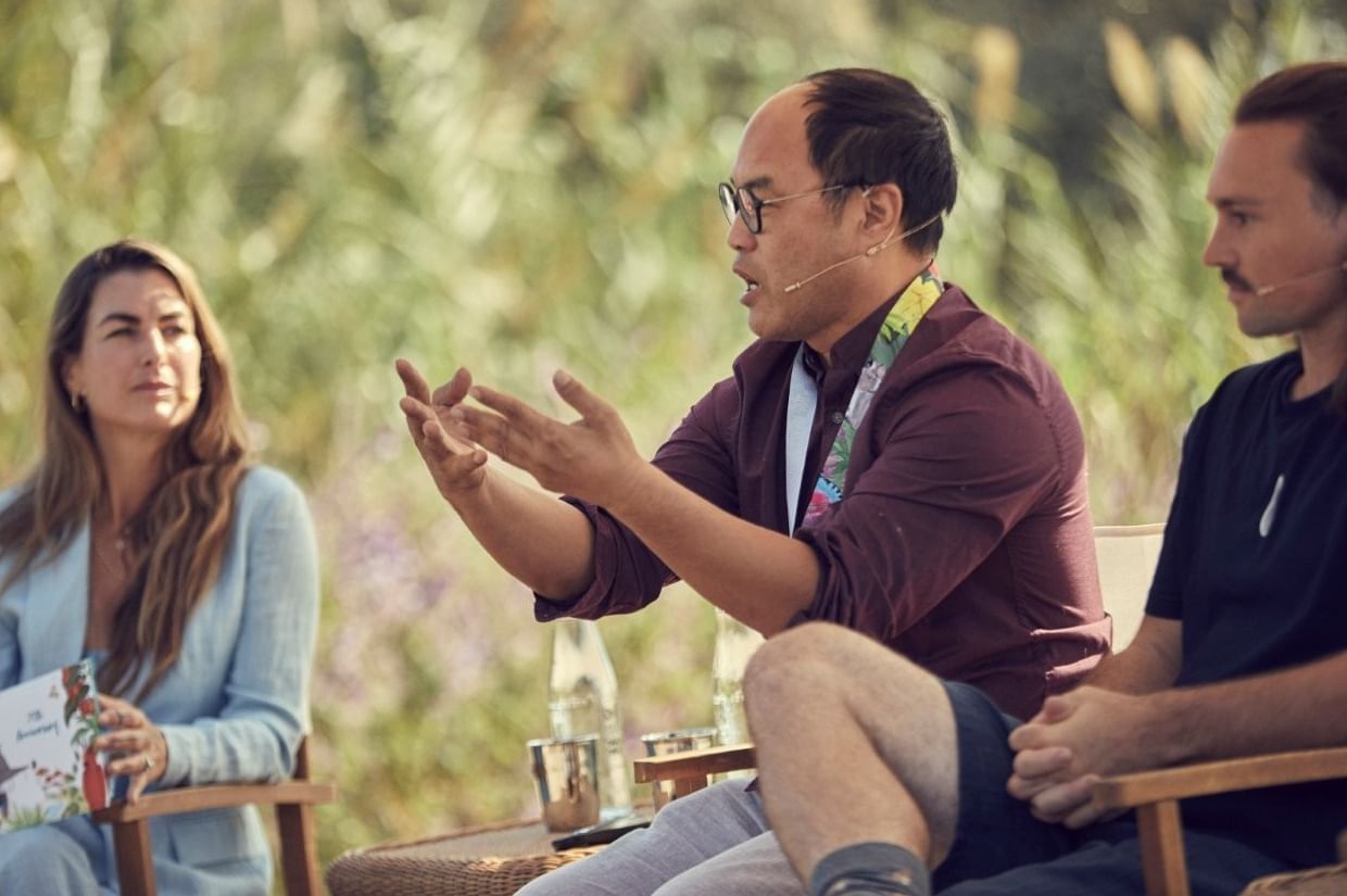 Woman speaking into a microphone and holding a program, with a blurred ocean backdrop near Marbella Club