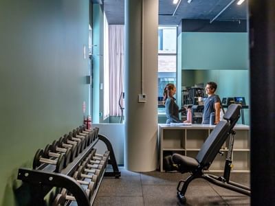 Modern fitness center with dumbbells, weight bench, and two residents conversing near the reception area at Swift Walk.
