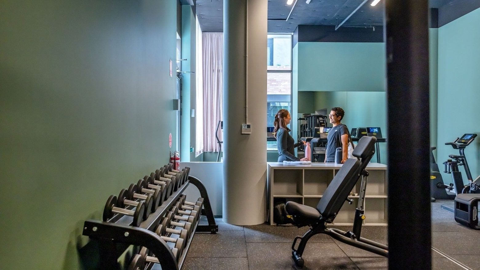 Modern fitness center with dumbbells, weight bench, and two residents conversing near the reception area at Swift Walk.
