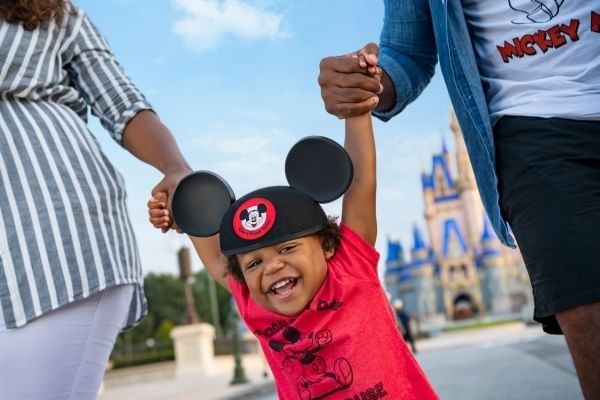 A laughing toddler in a red shirt and Mickey ears swings by the hands of two grown ups with Cinderella Castle in the background. Magic Kingdom is a great Disney park for toddlers.