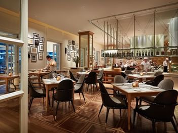 An opera interior with wooden tables, stylish chairs, and chefs working in an open kitchen at Park Hyatt Saigon
