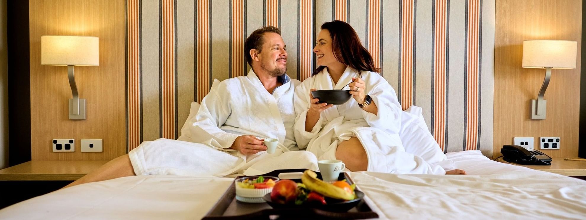 Couple in robes enjoying a tray of fruit and snacks on a hotel bed.
