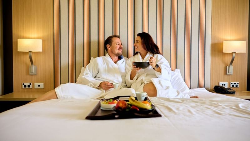 Couple in robes enjoying a tray of fruit and snacks on a hotel bed.