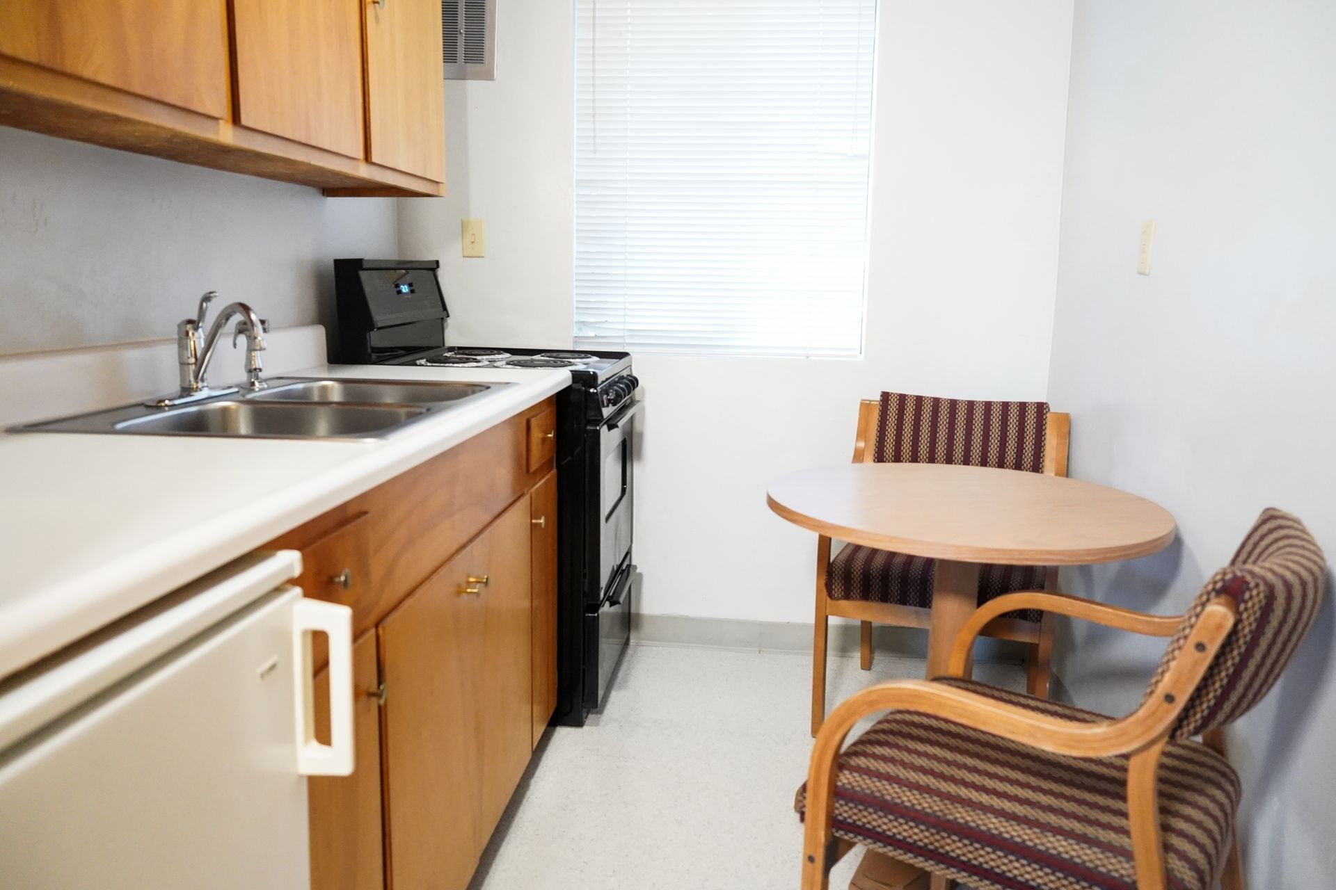 A compact kitchenette with wooden cabinets, a stainless steel sink, and a black stove, accompanied by a small dining area with a round table and two chairs.