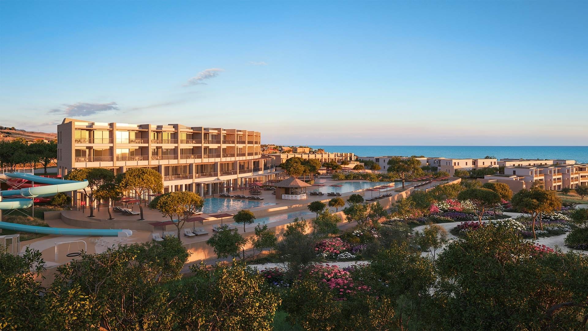Aerial view of resort with pool, slides, buildings, and ocean in background under a clear sky.