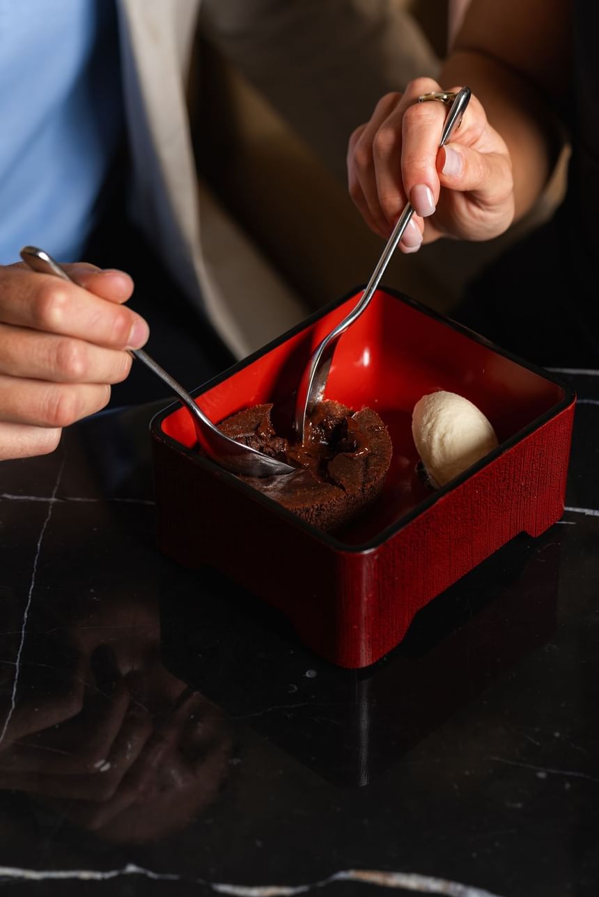 Two hands holding spoons dive into a rich chocolate lava cake served in a red bowl at The Kitchens