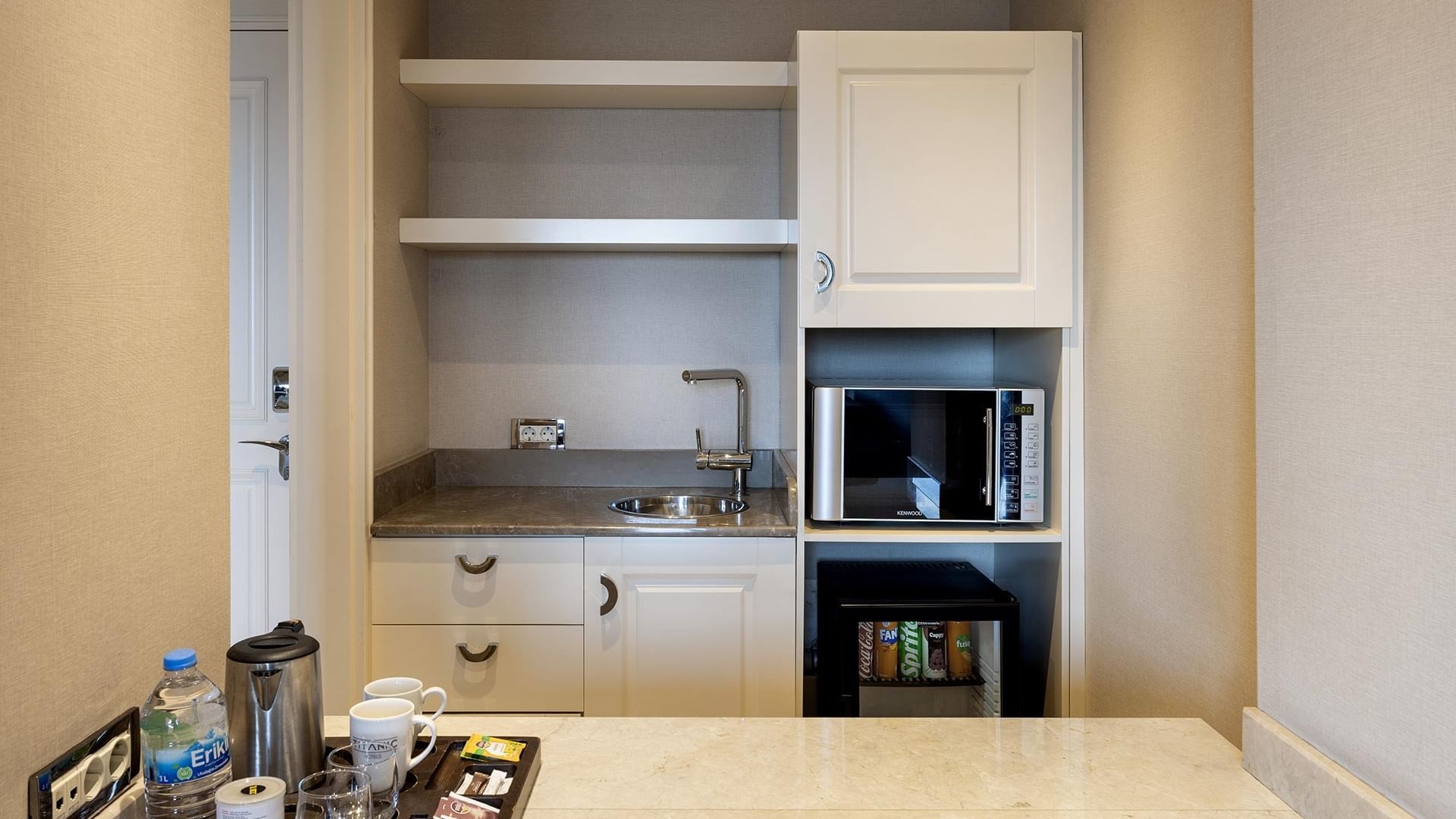 Compact kitchenette area with microwave, sink, mini bar, and tray with coffee, cups, and bottled water in the Titanic City Taksim hotel suite.