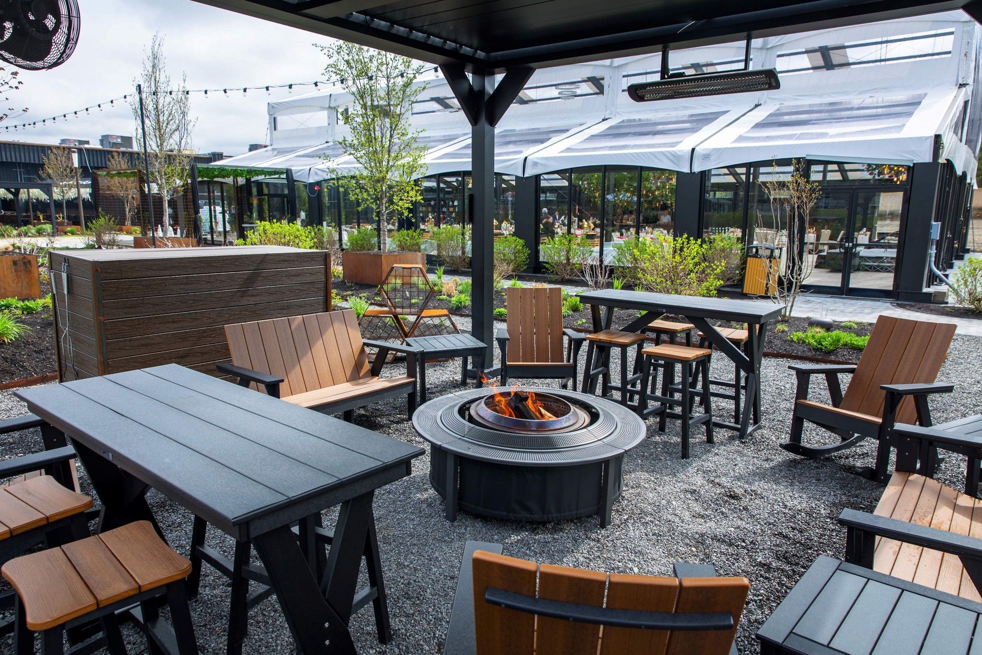 Wooden chairs surrounding a lit fire pit under a black canopy on a gravel patio in The Secret Garden Pergolas at The Brook