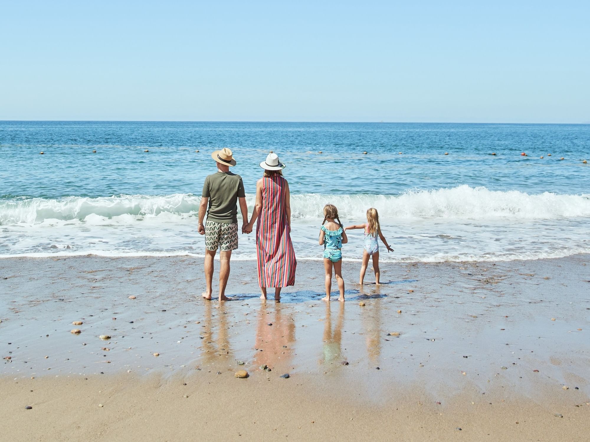 Familia de cuatro personas disfrutando de la orilla del mar bajo un cielo despejado en una playa de arena dorada.