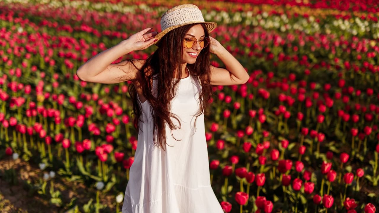 Woman in white dress and hat among red tulips, holding hat and smiling.
