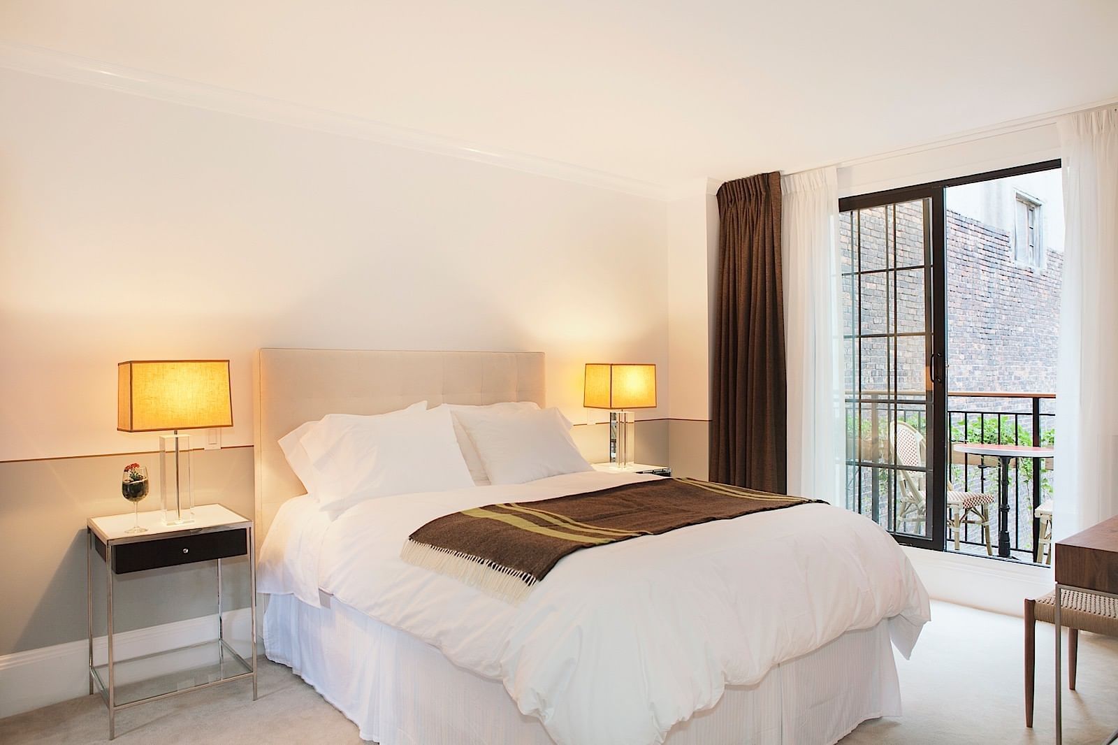 A neatly made bed with white linens and a brown throw blanket, two nightstands with lamps, and a large window leading to a balcony.
