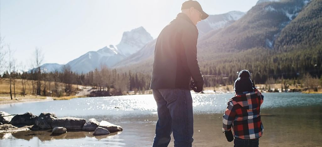 A grandparent and grandchild admire the view of the mountains near a river in Canmore.