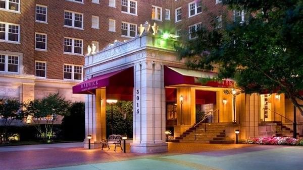 Red awnings by a stone entrance under glowing lights at the brick exterior of Warwick Melrose Dallas