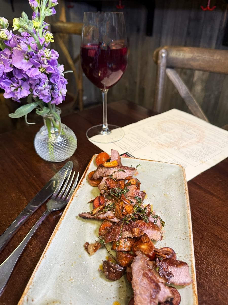 Sliced steak by purple flowers and a glass of wine on a dark table in BOTA at Warwick San Francisco