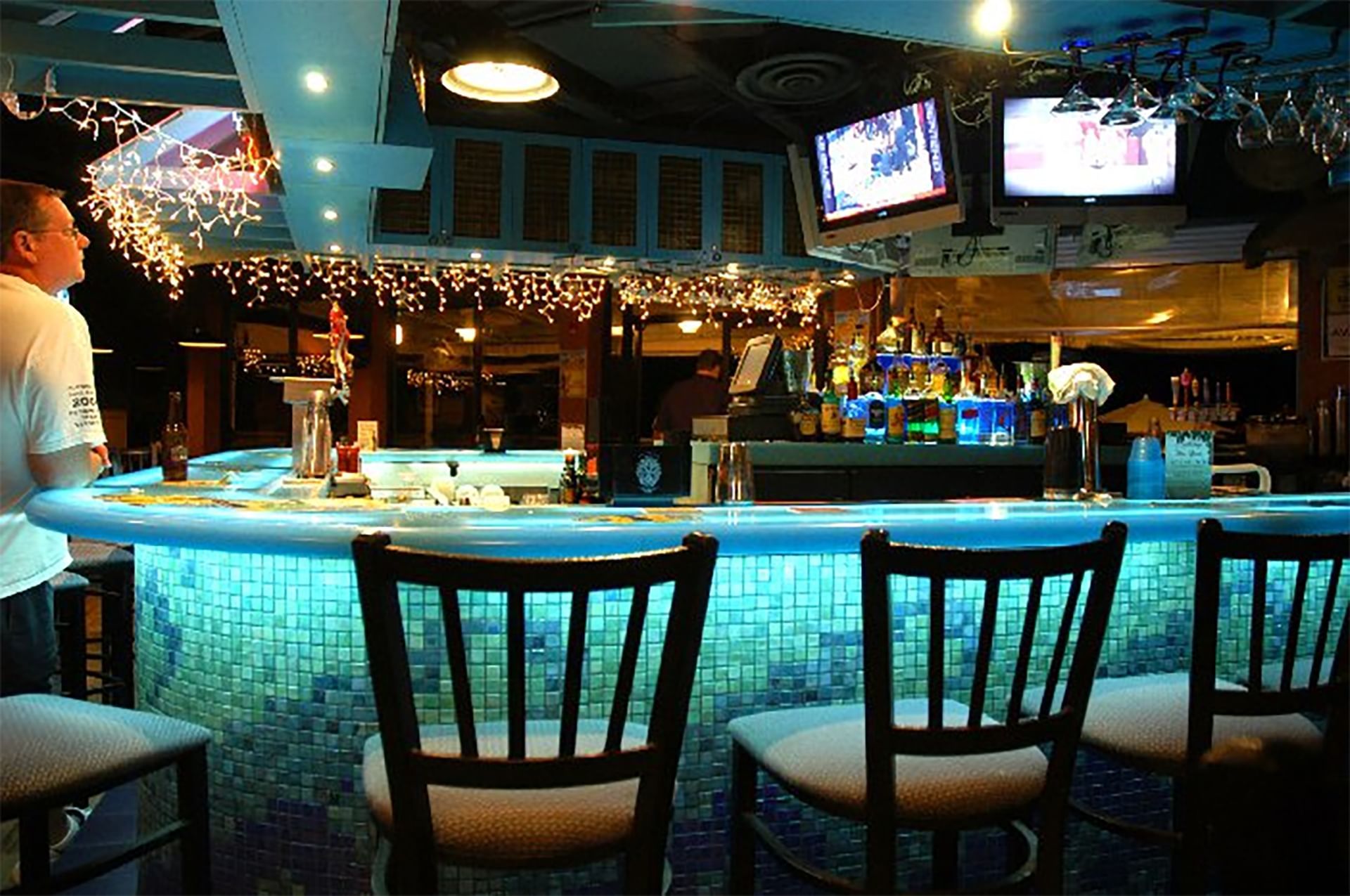 A dimly lit bar with a curved, tiled counter and stools, featuring glowing blue lights, TVs, and string lights.