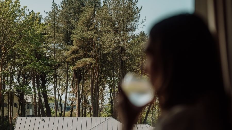 Guest holding a drink by the window overlooking tall green pine trees and peaceful surroundings at Dunluce Lodge