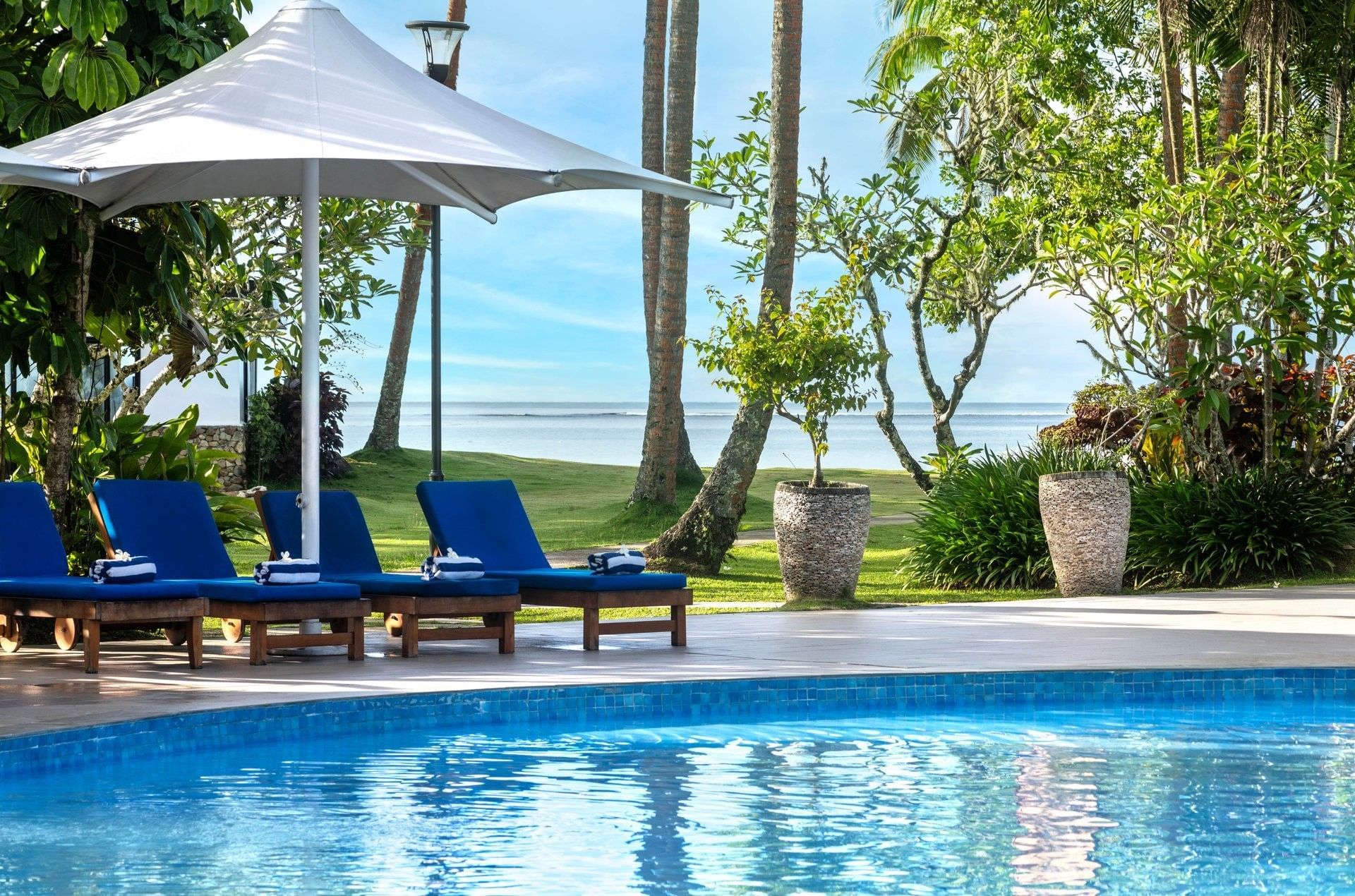 Blue sunbeds under a white umbrella by a pool near the ocean and palm trees at The Naviti Resort - Fiji