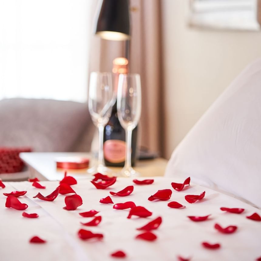 Pétalos de rosa sobre una cama blanca junto a copas de champán y una pequeña caja de regalo en Megapolis Hotel Panama