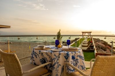 An intimate candlelit dinner table setup for two directly on the sand against a stunning ocean sunset.